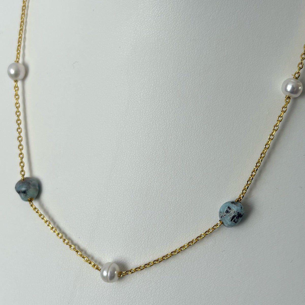 Close-up of white baroque pearls alternating with blue-grey larimar stones on a gold-filled chain, displayed on a white jewellery bust, showing the stones’ dark markings.⠋