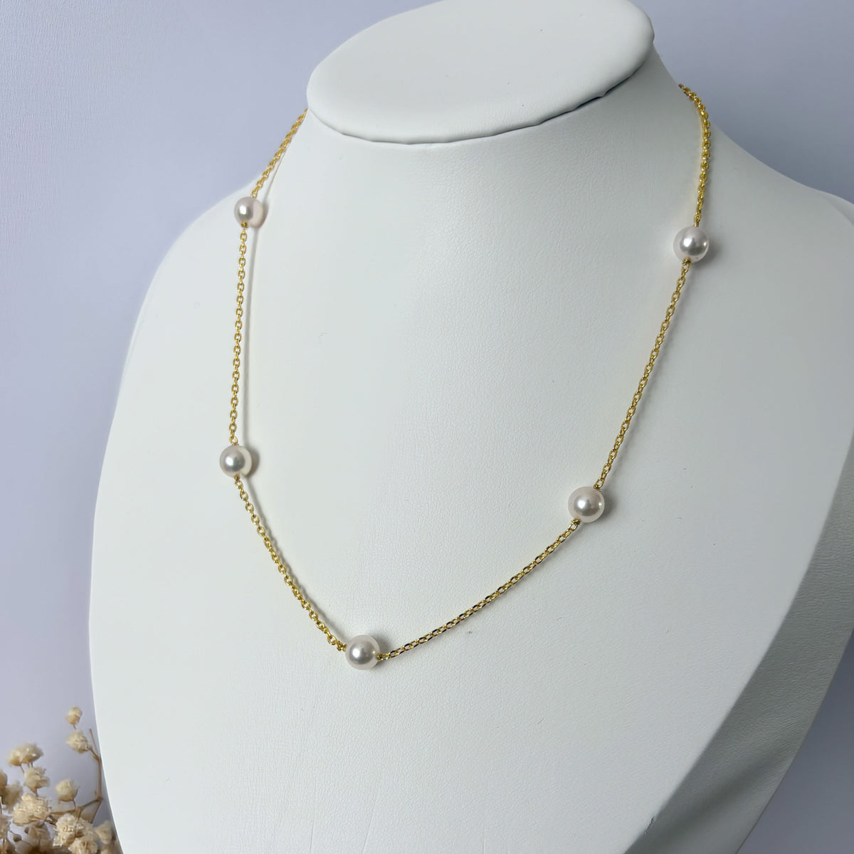 Five white Akoya pearls spaced along a gold-filled chain, displayed on an angled white jewellery bust with dried flowers at its base.