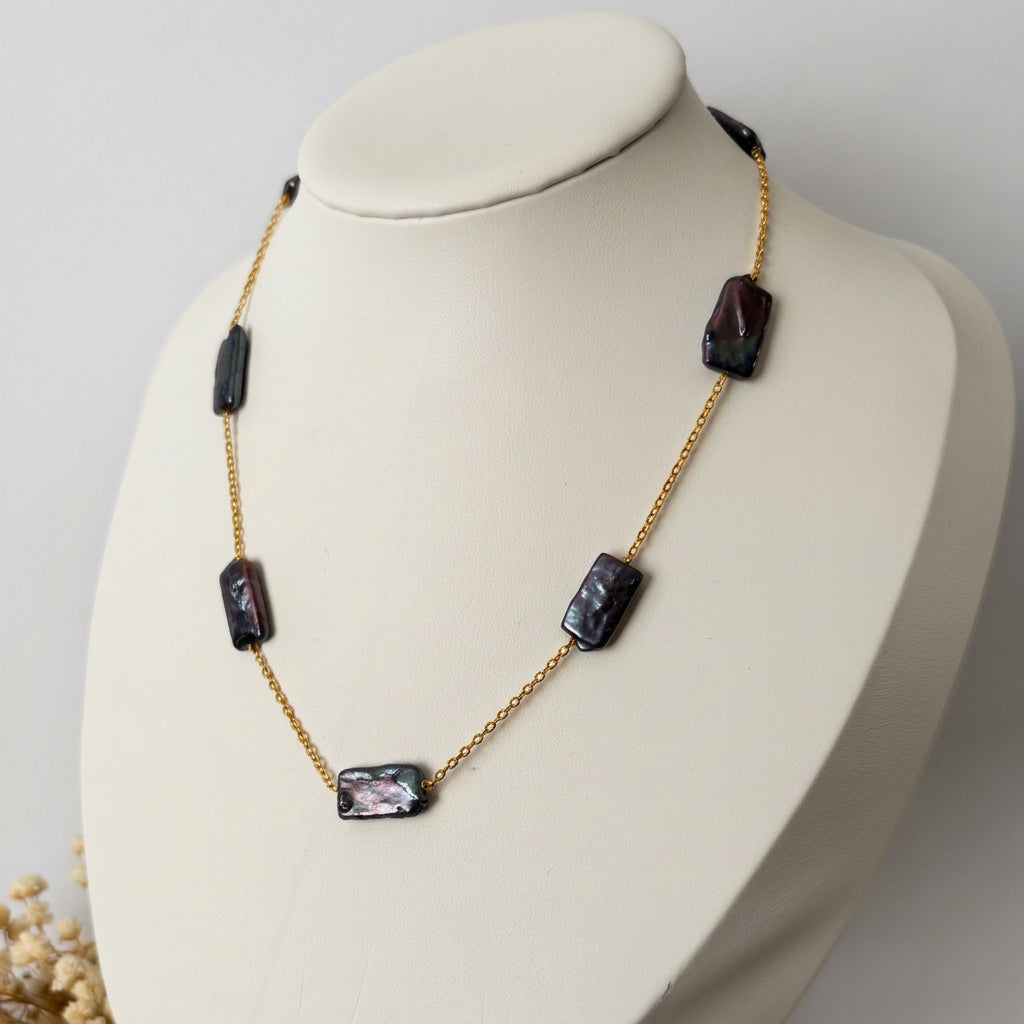 Dark blue rectangular baroque pearls spaced along a gold-filled chain, displayed on an angled white jewellery bust with dried flowers at its base.