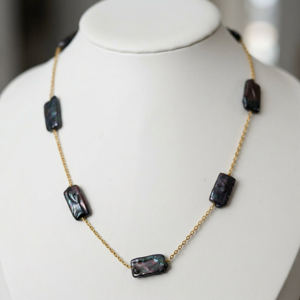 Dark blue rectangular baroque pearls spaced along a fine gold-filled chain, displayed on a white jewellery bust, showing their irregular surfaces and purple-green iridescence.