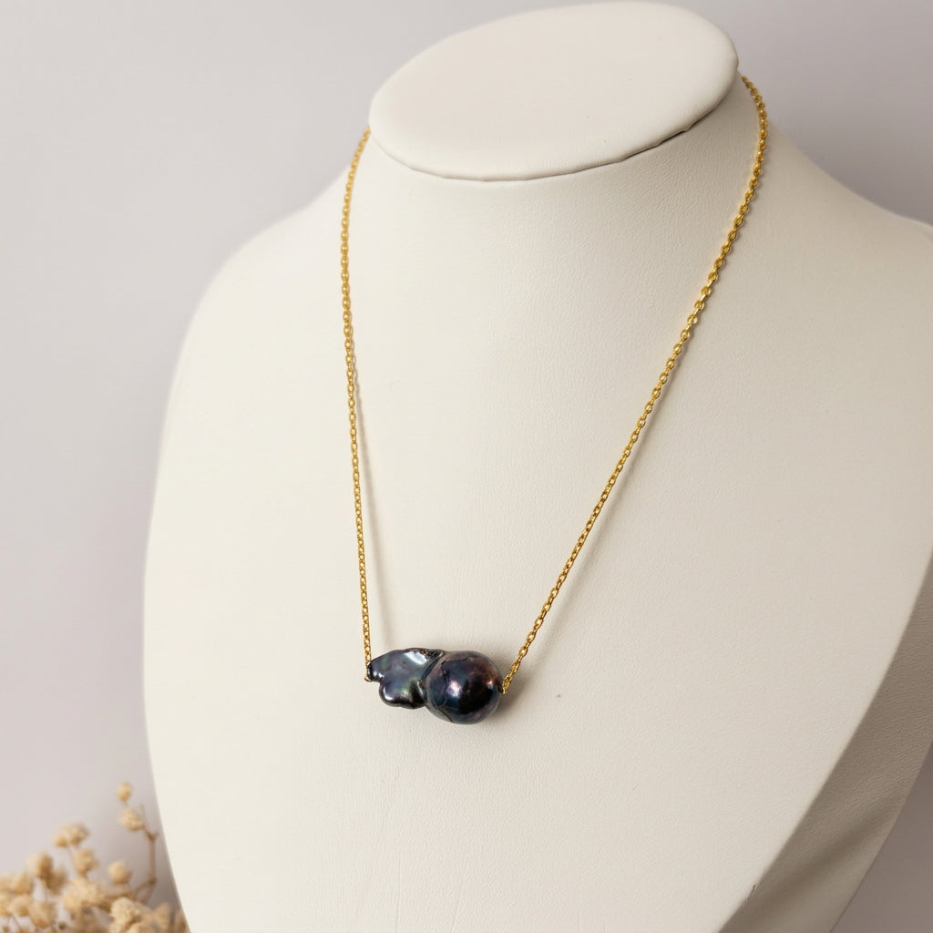 Dark blue keshi pearl on a fine gold-filled chain, displayed on a white jewellery bust beside dried flowers, showing the pearl’s rounded dome and irregular folded shape.