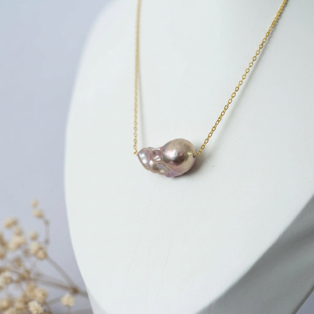 Rainbow keshi pearl on a fine gold-filled chain, displayed on a white jewellery bust beside dried flowers, showing the pearl’s irregular shape and champagne-pink lustre.