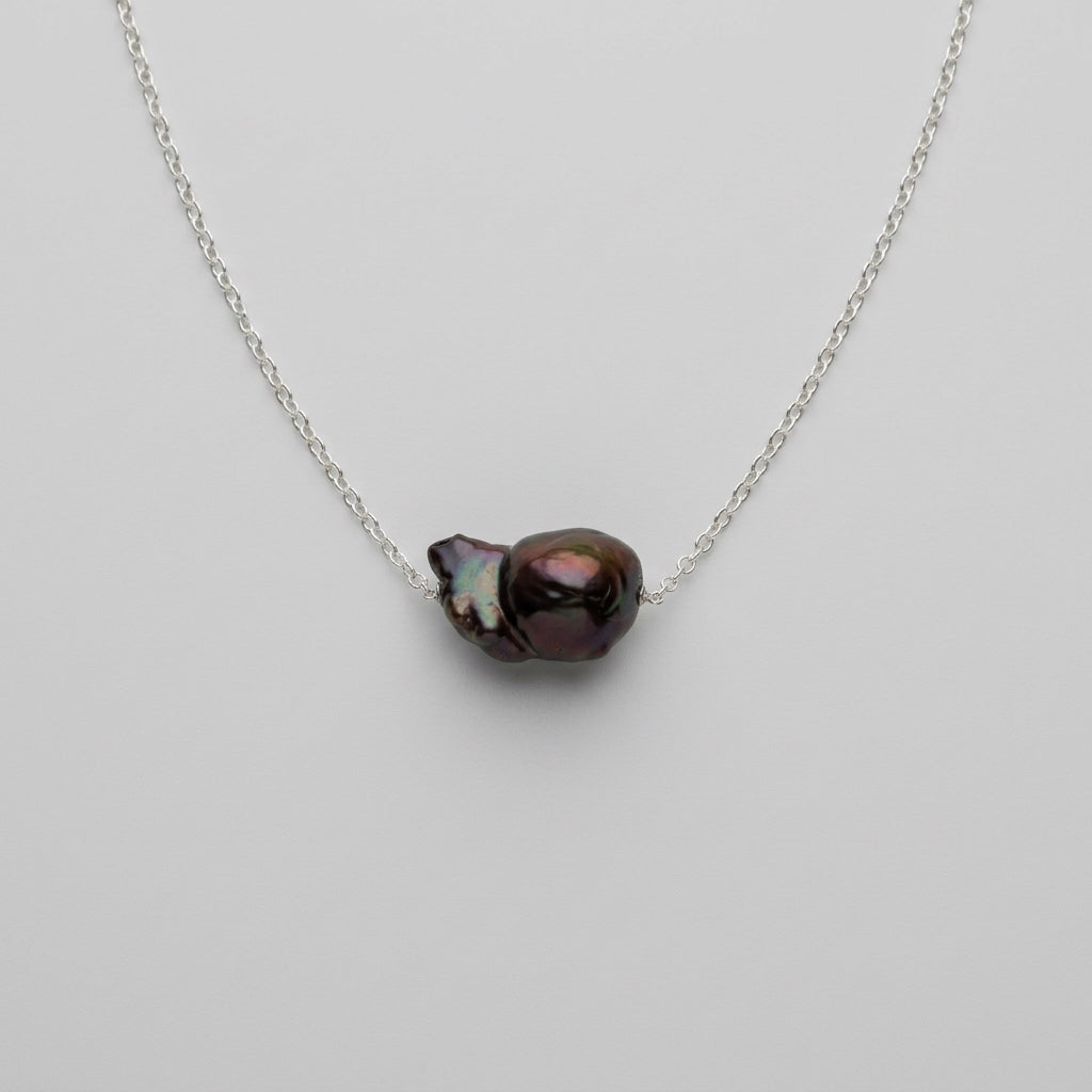 Close-up of a dark blue Keshi pearl on a sterling silver chain against a pale grey background, showing its irregular shape and purple-green iridescent highlights.