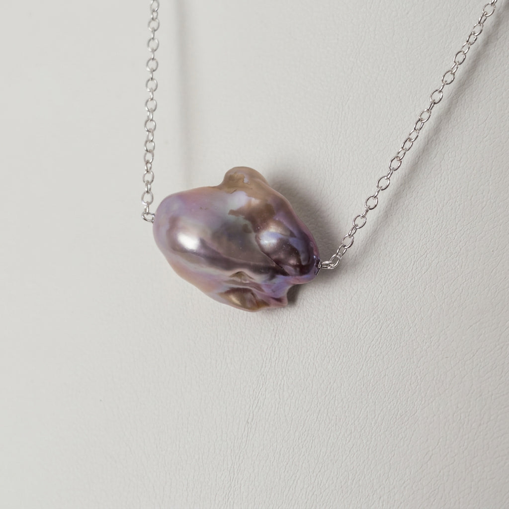 Close-up of a pink Keshi pearl on a fine chain against a white jewellery bust, showing its irregular folds, rounded contours and mauve-pink iridescence.