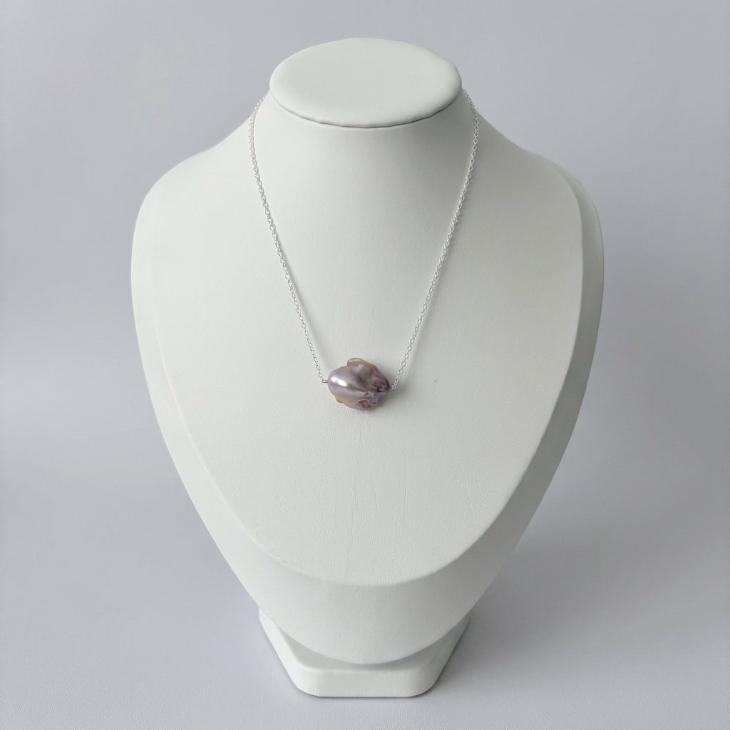 Close-up of a lavender Keshi pearl set on a sterling silver chain against a pale grey background, showing its irregular contours and lustrous purple and silver tones.