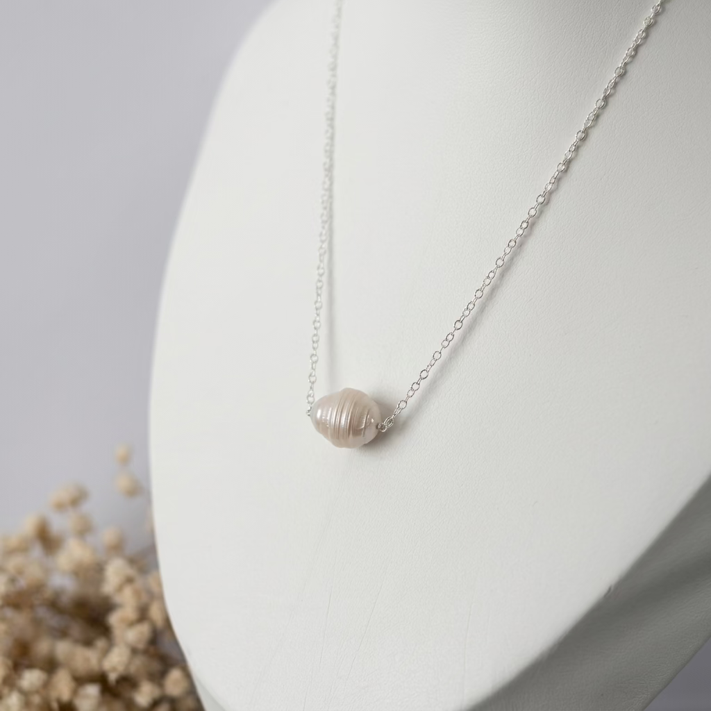 White ribbed baroque pearl necklace on a fine sterling silver chain displayed on a white jewellery bust, with dried cream flowers beside the display