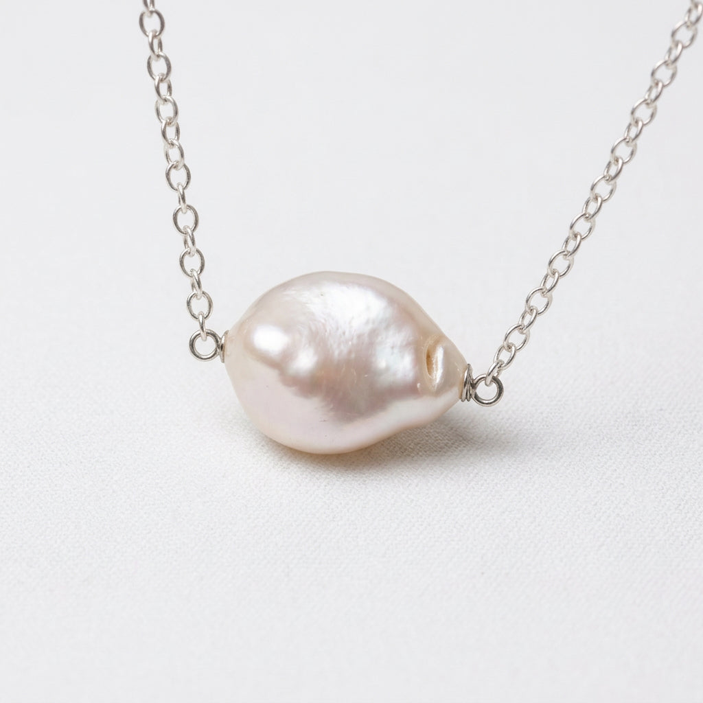 White baroque pearl necklace on a fine sterling silver chain displayed on a white jewellery bust, with dried cream flowers beside the display.