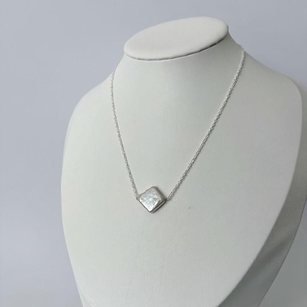 White square baroque pearl set diagonally on a fine sterling silver chain, displayed on an angled white jewellery bust against a pale grey background.