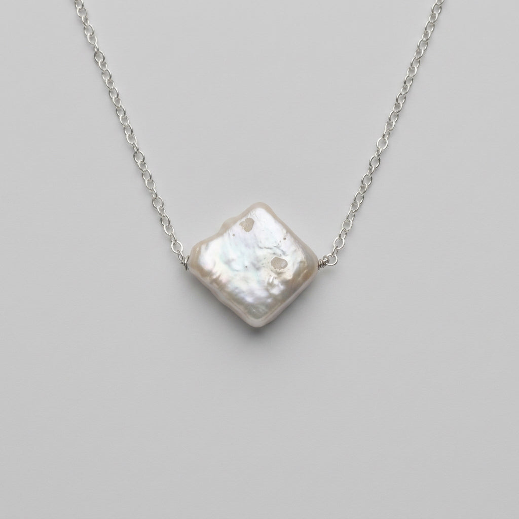 Close-up of a white square baroque pearl set diagonally on a sterling silver chain against a pale grey background, showing its irregular surface and iridescent sheen.⠋