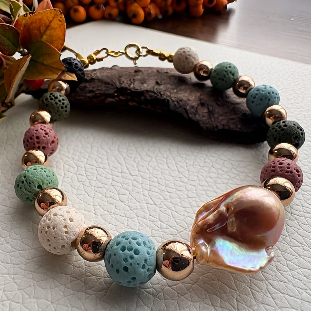 Multicoloured lava bead bracelet with polished gold-plated spacers and a large peach-gold keshi pearl, arranged beside bark and autumn leaves on a white textured surface.