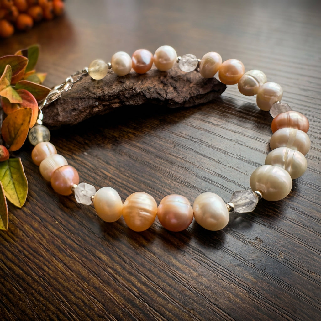 Creamy white and peach baroque pearl bracelet with faceted clear quartz, pale yellow citrine and sterling silver spacers, arranged on a wooden surface beside bark and autumn leaves.