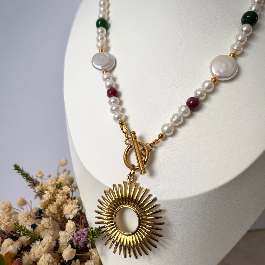 White baroque and coin pearl necklace displayed on a white bust beside dried flowers, with red and green tourmaline beads, a fastened gold-plated toggle clasp and an open-circle sunburst pendant.