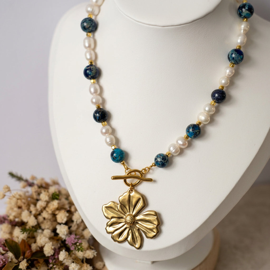 Blue jasper and white baroque pearl necklace displayed on a white bust beside dried flowers, with gold-coloured hematite spacers, a fastened front toggle clasp and a large gold-plated flower pendant.
