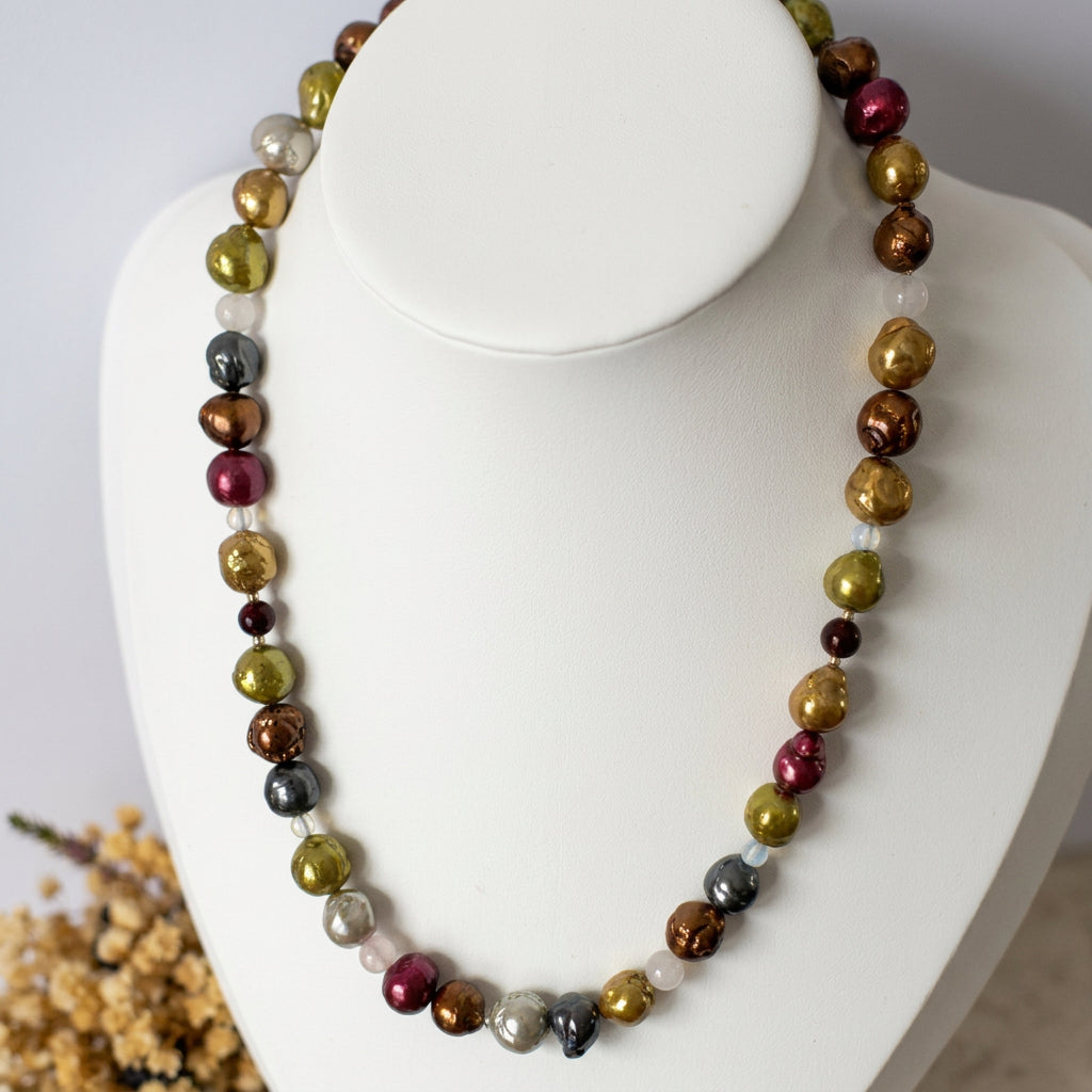 Multicoloured baroque pearl necklace displayed on a white bust beside dried flowers, showing gold, lime-green, bronze, burgundy and silver-grey pearls interspersed with pale gemstone accents and dark red garnet beads.⠋
