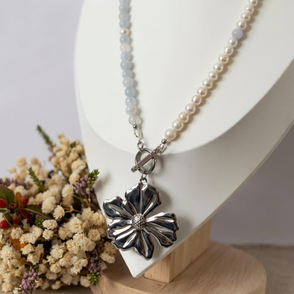 White baroque pearl and pale blue aquamarine necklace with a front toggle clasp and large silver-coloured flower pendant, displayed on a white jewellery bust beside dried flowers.