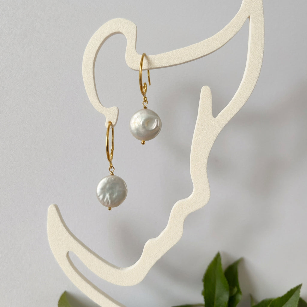 Pair of white coin pearl drop earrings with gold-filled hooks and small gold bead tips, hanging from a cream face-shaped display.