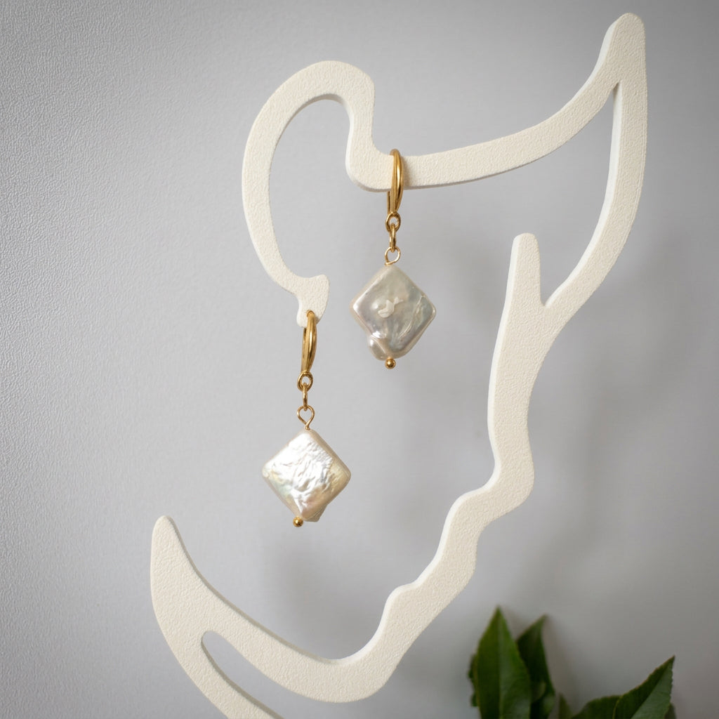 Pair of white square baroque pearl drop earrings with gold-filled hooks, hanging diagonally from a cream face-shaped display and showing textured, lustrous surfaces.