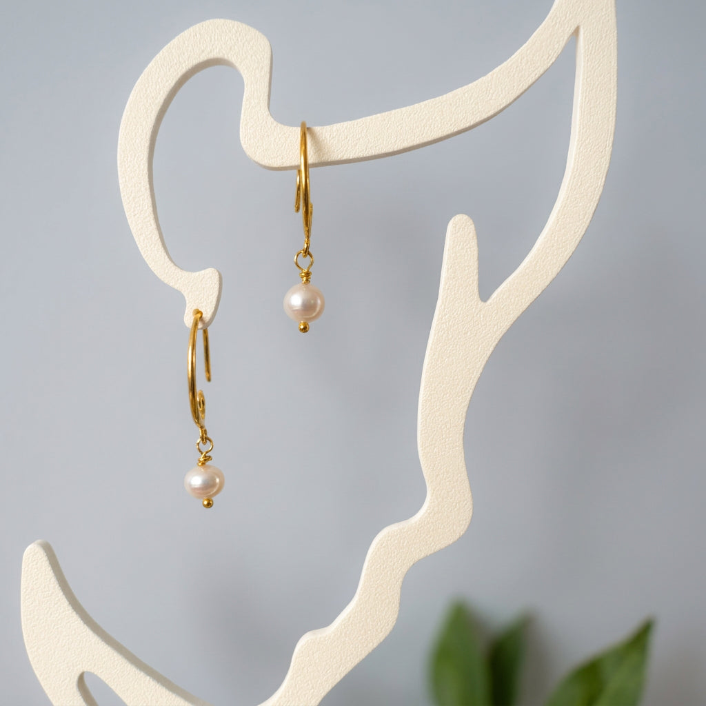 Pair of white baroque pearl drop earrings with curved gold-filled hooks and small gold bead tips, hanging from a cream face-shaped display.