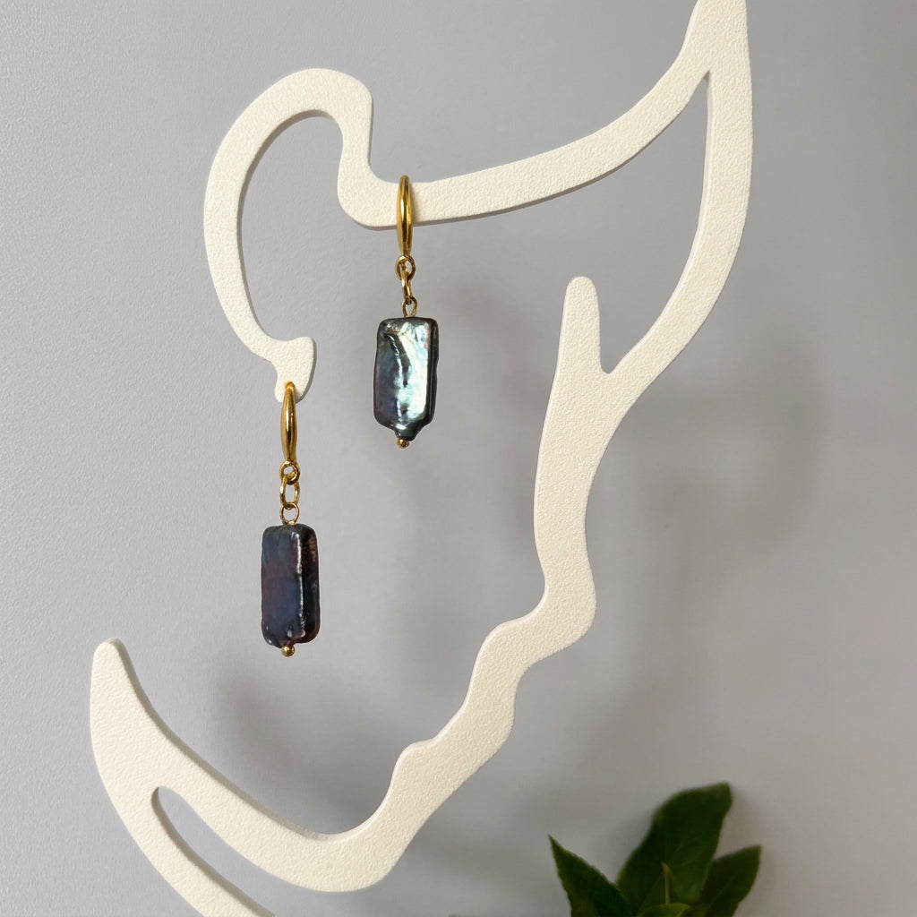 Pair of dark blue square baroque pearl drop earrings with gold-filled hooks, hanging from a cream face-shaped display and catching silvery-blue highlights.