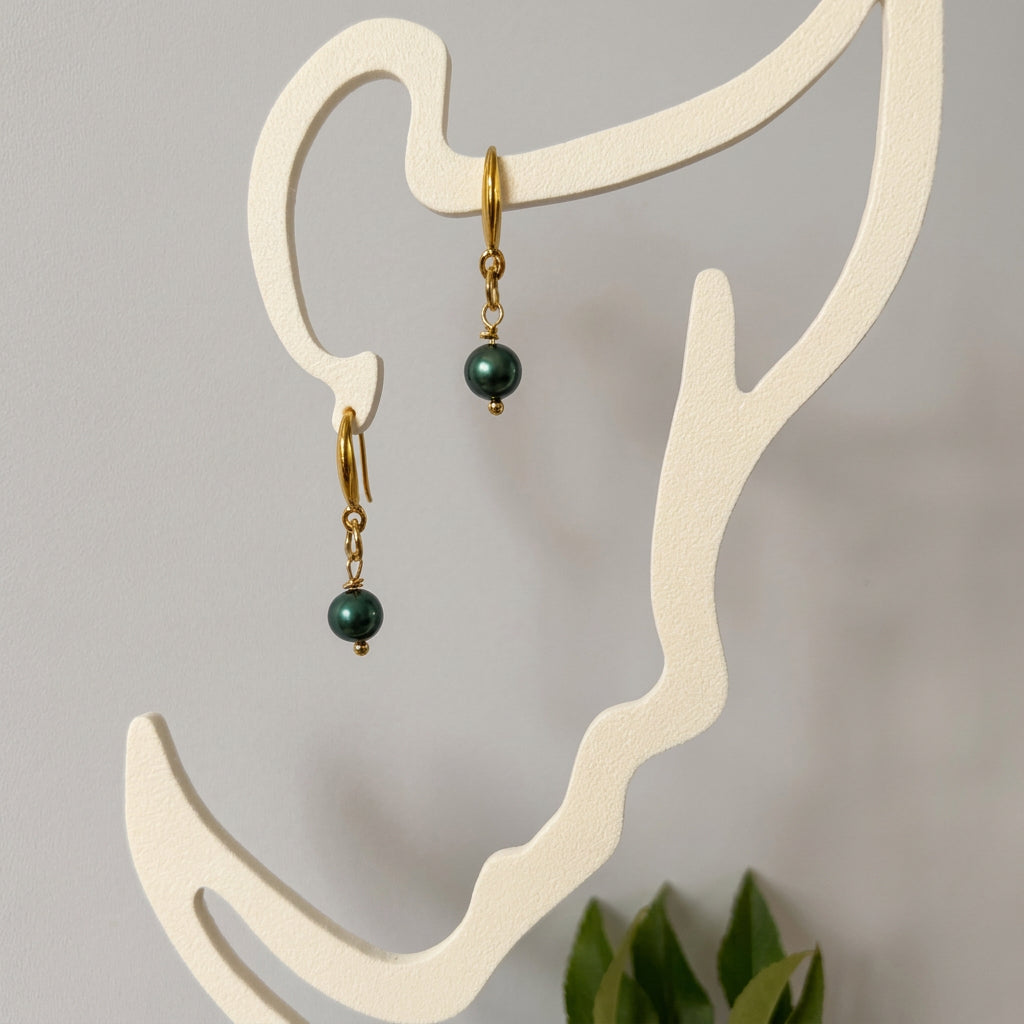 Pair of deep green peacock baroque pearl drop earrings with gold-filled hooks and twisted links, hanging from a cream face-shaped display.