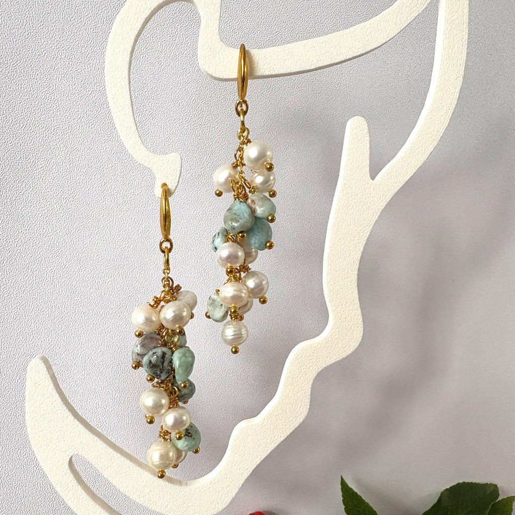 Pair of long cluster drop earrings with white baroque pearls and pale blue-green stones, hanging from gold-filled hooks on a cream face-shaped display.
