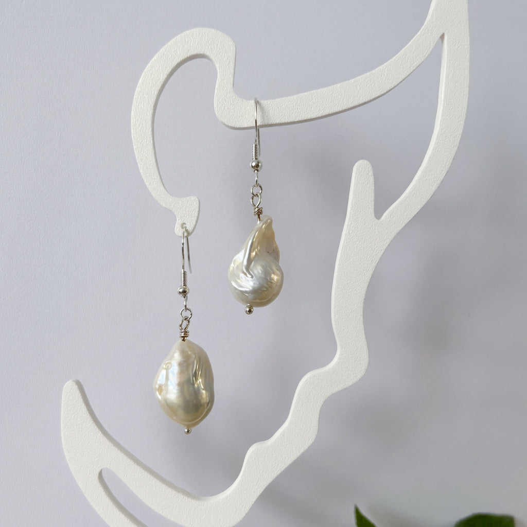 Pair of white Keshi pearl drop earrings with irregular elongated shapes and sterling silver hooks, hanging from a cream face-shaped display.