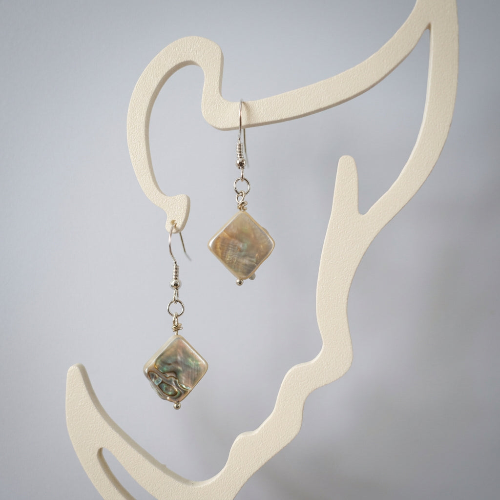 Pair of white square baroque pearl drop earrings with sterling silver hooks, hanging diagonally from a cream face-shaped display and showing their textured nacre surfaces.