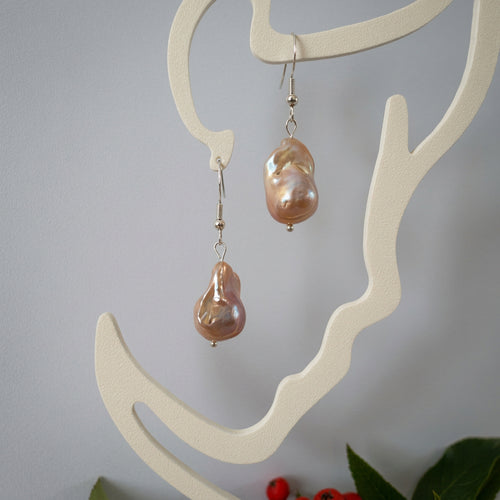 Pair of cream keshi pearl drop earrings displayed on a white branching stand, showing irregular golden-peach pearls with iridescent highlights suspended from sterling silver hooks.⠋