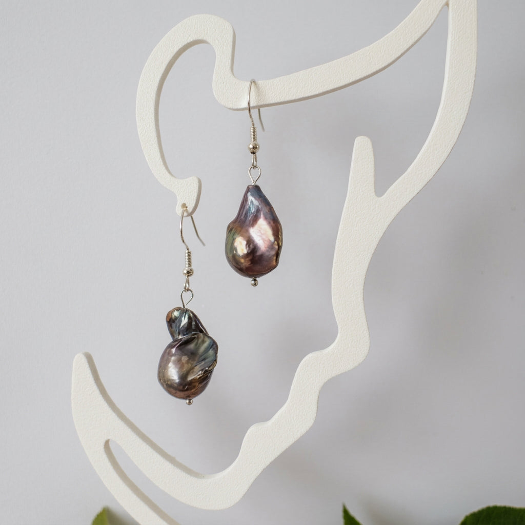 Pair of irregular dark keshi pearl earrings on sterling silver hooks, hanging from a white display stand with pink, bronze and silver iridescent highlights.