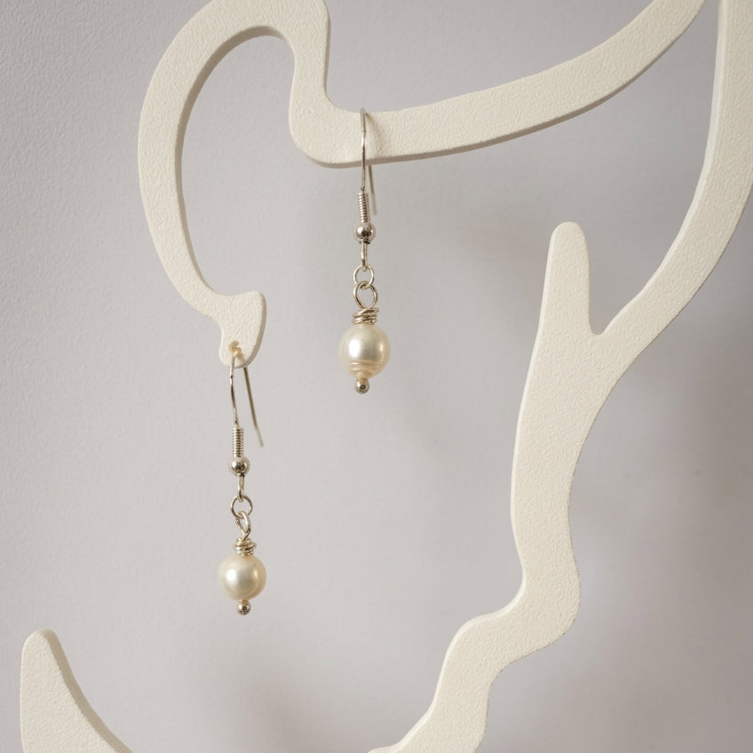 Pair of white baroque pearl drop earrings with sterling silver hooks and connecting links, hanging from a cream face-shaped display.