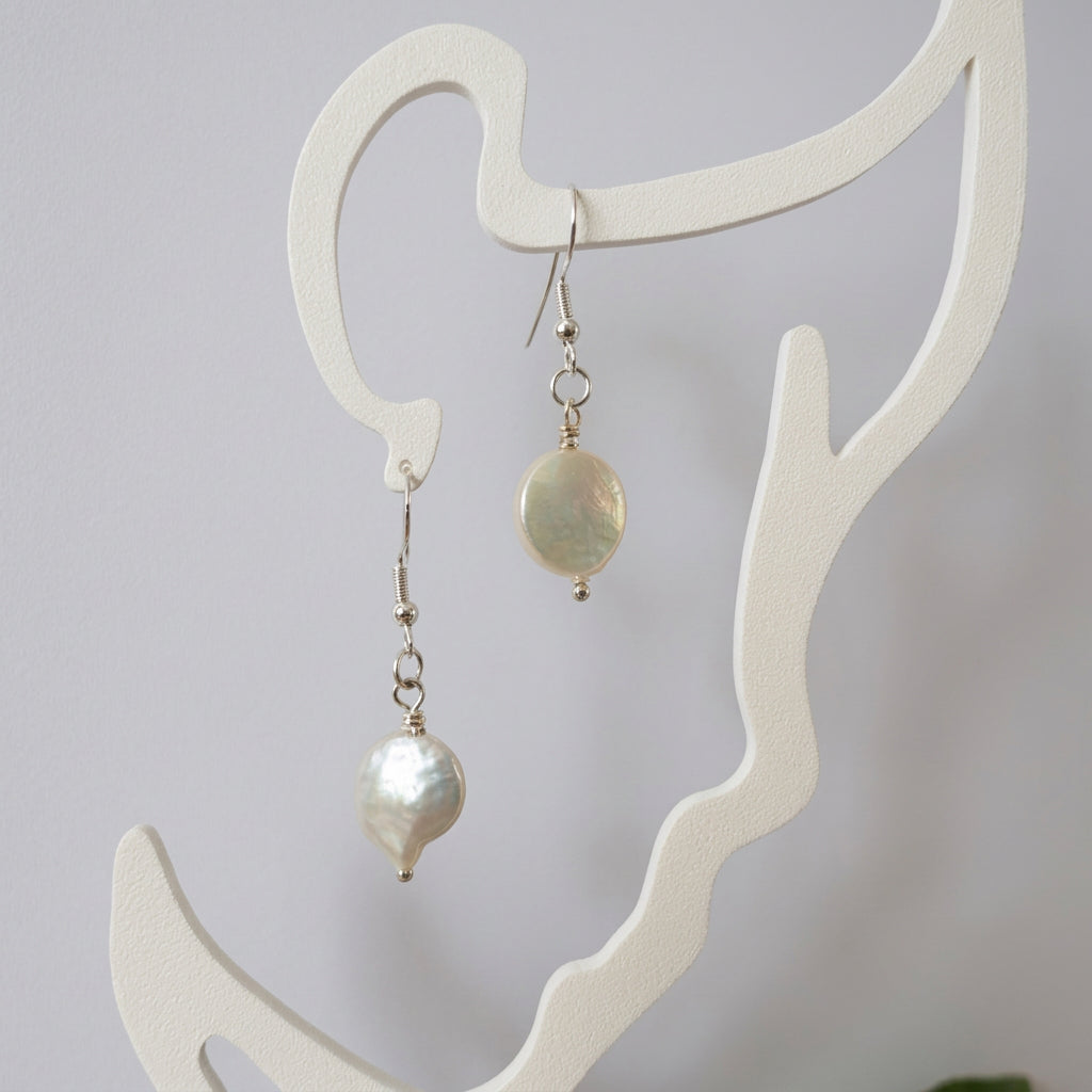 Pair of white coin pearl drop earrings with sterling silver hooks and connecting rings, hanging from a cream face-shaped display.⠋