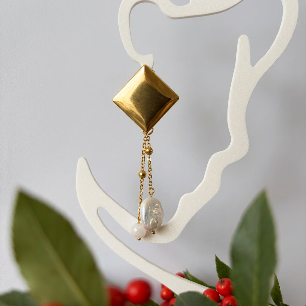 Gold-coated diamond-stud drop earring displayed on a white outline face stand above green leaves and red berries, with twin beaded chains ending in an oval white baroque pearl and a pale pink quartz bead.