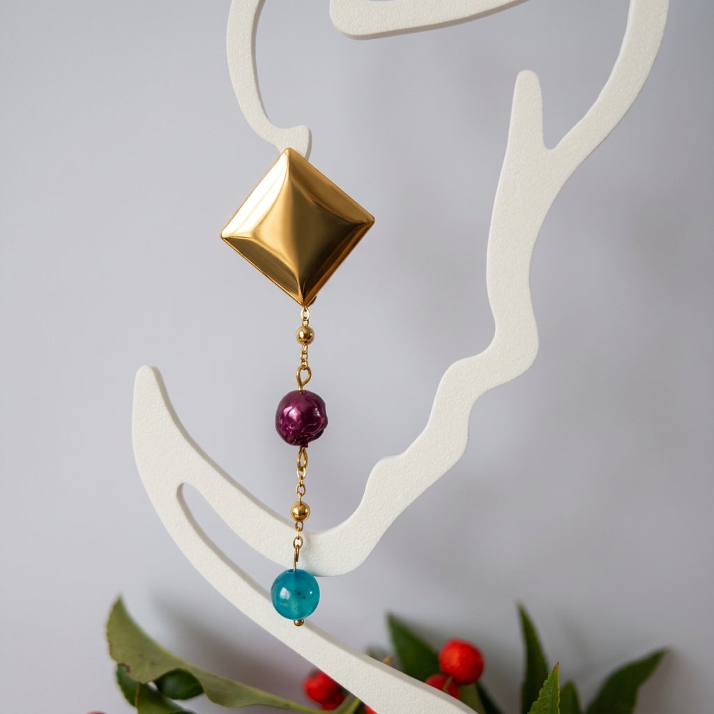 Gold-coated diamond-stud drop earring displayed on a white outline face stand, with a burgundy baroque pearl and bright blue topaz bead along a fine beaded chain, above green leaves and red berries.⠋
