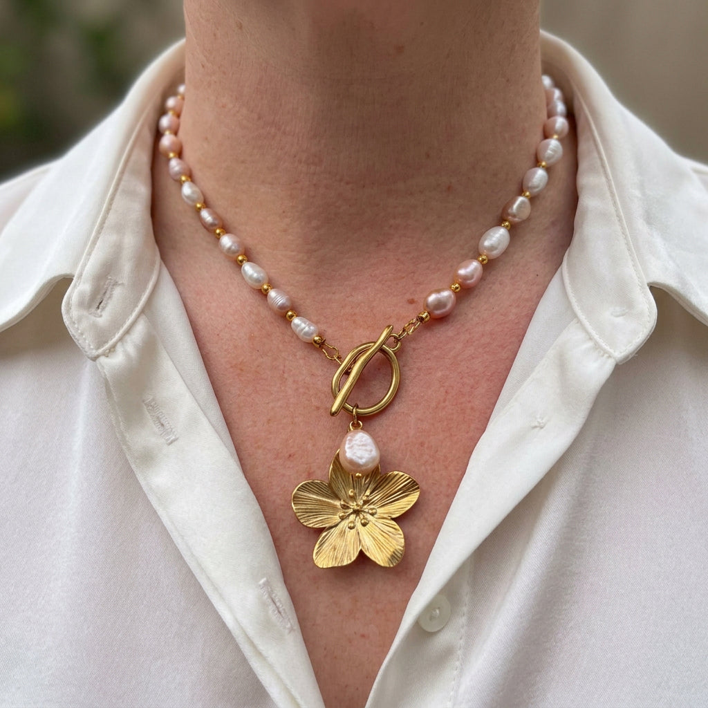 White and blush-pink baroque pearl necklace with gold-coloured spacers, a fastened front toggle, a white pearl drop and a textured four-petal gold-coloured flower pendant, worn with an open-neck white shirt.