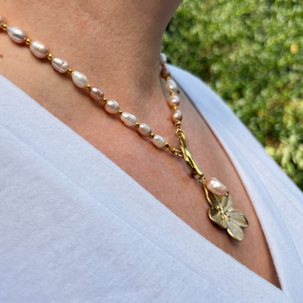 Side-angle view of a white and blush pink baroque pearl necklace with gold-plated spacer beads, a front toggle clasp, pearl drop and gold-coloured flower pendant, worn with a white V-neck top outdoors.⠋