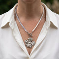 White baroque pearl and pale blue aquamarine necklace with a fastened front toggle clasp and large silver-coloured sculpted flower pendant, worn with an open-neck white shirt.