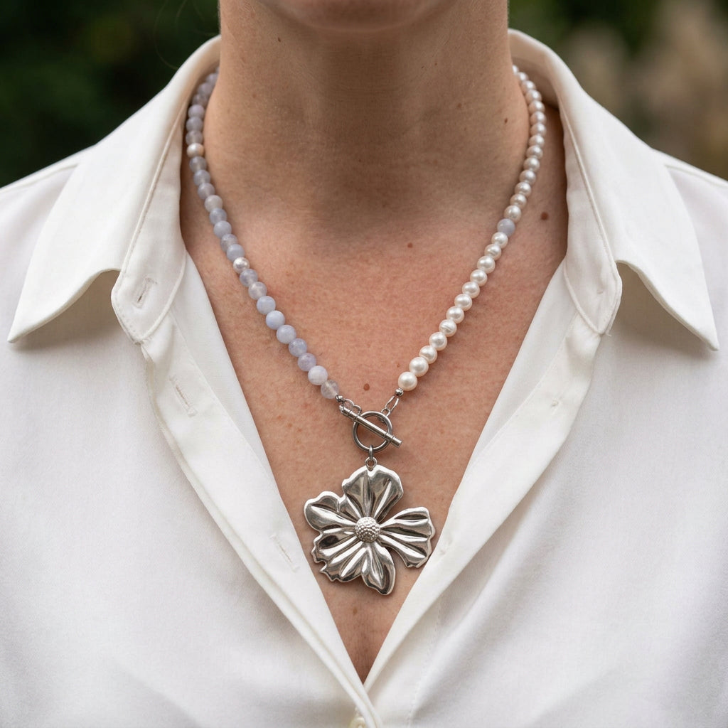 White baroque pearl and pale blue aquamarine necklace with a fastened front toggle clasp and large silver-coloured sculpted flower pendant, worn with an open-neck white shirt.