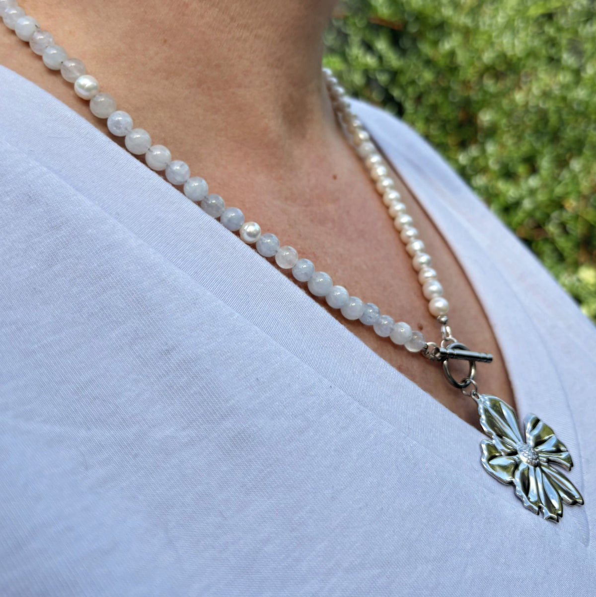 Side-angle view of a pale blue aquamarine and white baroque pearl necklace with a front toggle clasp and silver-coloured flower pendant, worn over a white V-neck top.