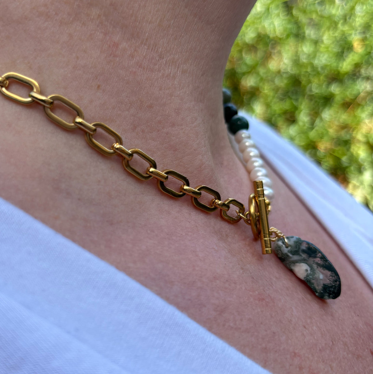 Side-angle view of a gold-plated paperclip chain necklace with white baroque pearls, green obsidian beads and a mottled green Indian agate pendant, worn with a white V-neck top outdoors.