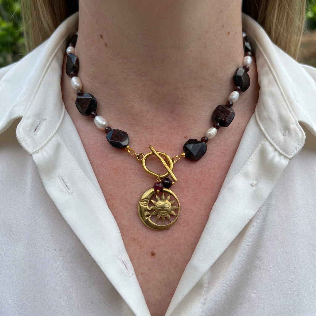 Deep burgundy garnet stones alternating with white rice pearls, with a fastened gold-plated front toggle clasp, two garnet drops and a sun-and-crescent-moon pendant, worn with an open-neck white shirt.