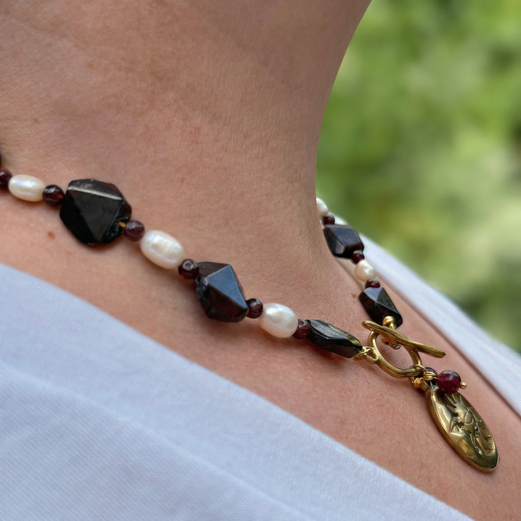 Side-angle view of a deep red faceted garnet and white rice pearl necklace with a gold-plated front toggle clasp and sun-and-crescent-moon pendant, worn with a white V-neck top outdoors.