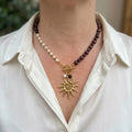 Burgundy garnet and white rice pearl necklace with a fastened gold-plated front toggle, pearl and garnet drops, and a large spiral-centred sun pendant, worn with an open-neck white shirt.