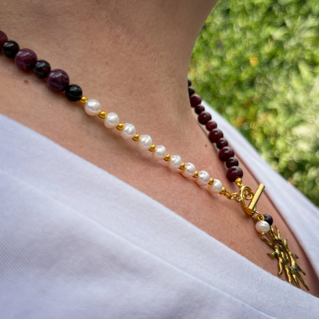 Side-angle view of a burgundy garnet and white baroque pearl necklace with gold-plated spacer beads, a front toggle clasp and gold-coloured sun pendant, worn with a white V-neck top outdoors.