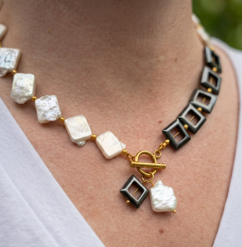 Angled close-up of a half-pearl, half-hematite necklace worn with a white V-neck top, showing rectangular white baroque pearls, black open-square beads and matching pendants beneath a gold-plated toggle clasp.