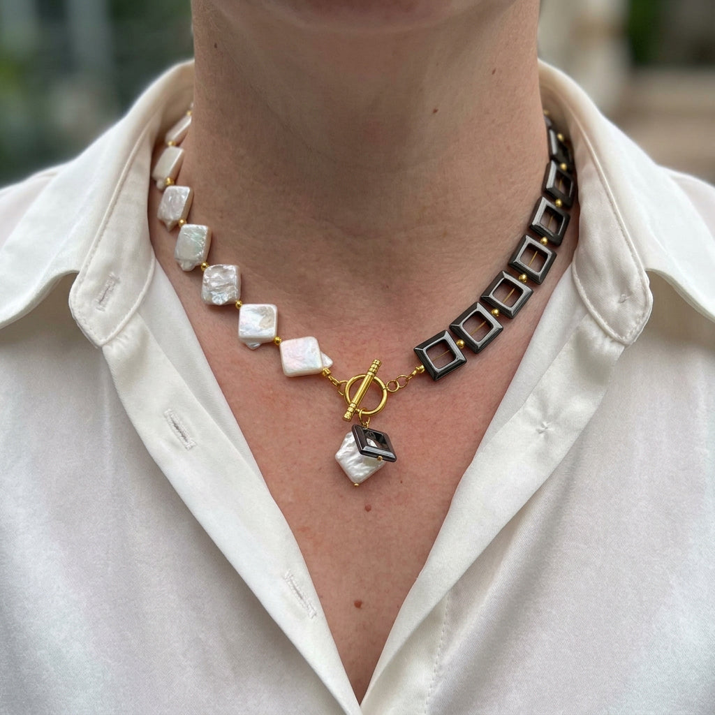 Half white square baroque pearl and half black open-square hematite necklace, with a fastened gold-plated front toggle and matching pearl and hematite drops, worn with an open-neck white shirt.