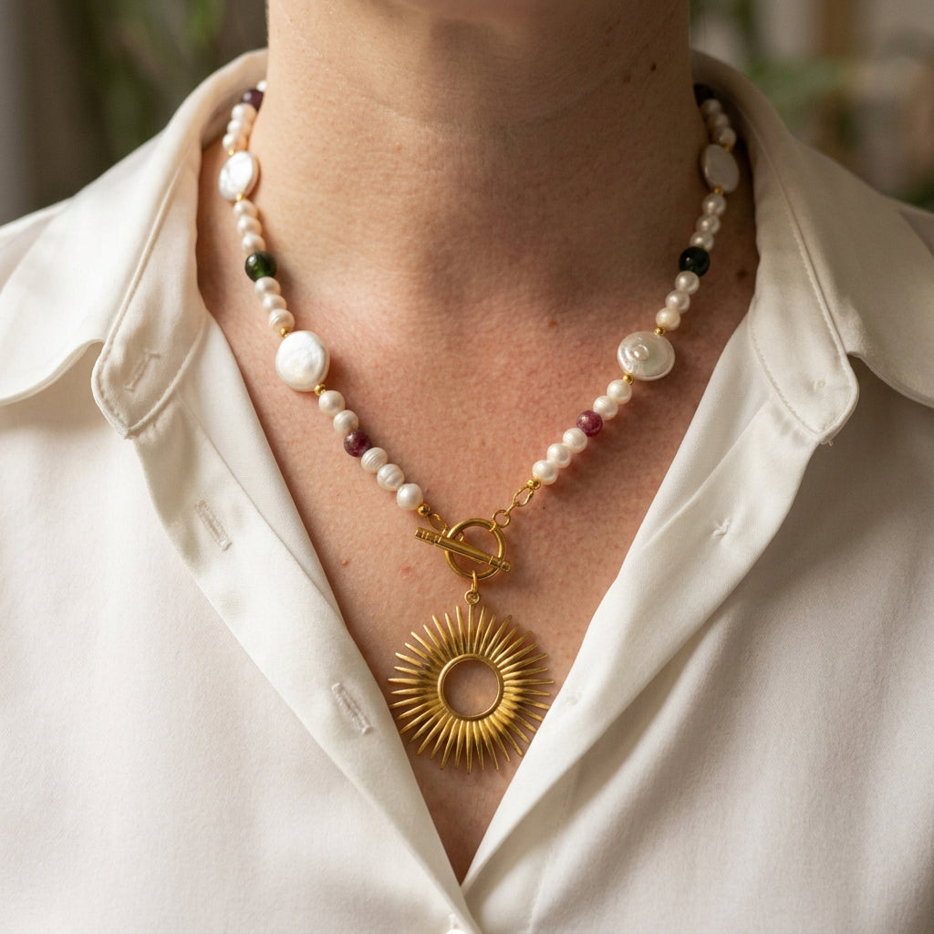 White baroque and coin pearl necklace with red and green tourmaline beads, a fastened gold-plated front toggle and large open-circle sunburst pendant, worn with an open-neck white shirt.