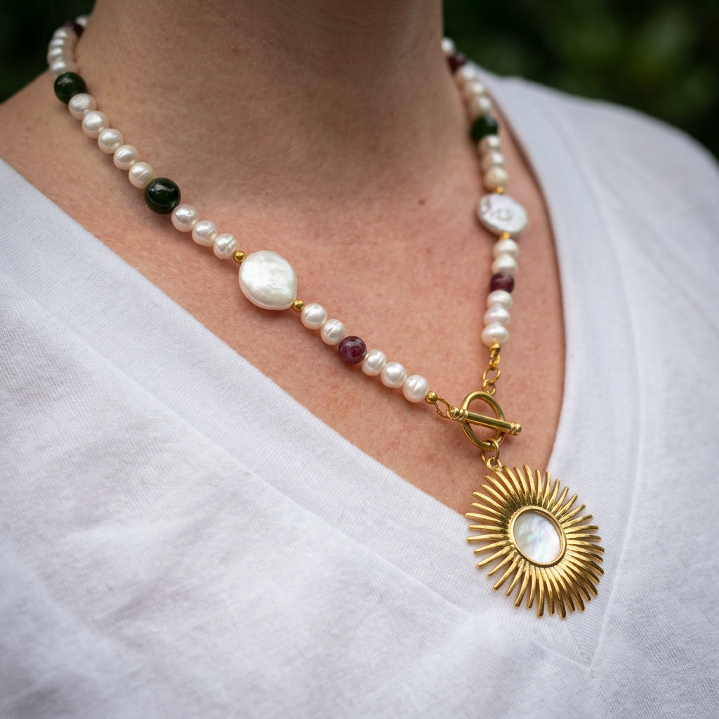 Angled view of a white baroque and coin pearl necklace worn with a white V-neck top, with red and green tourmaline beads, a fastened front toggle clasp and a gold-plated open-circle sunburst pendant.⠋