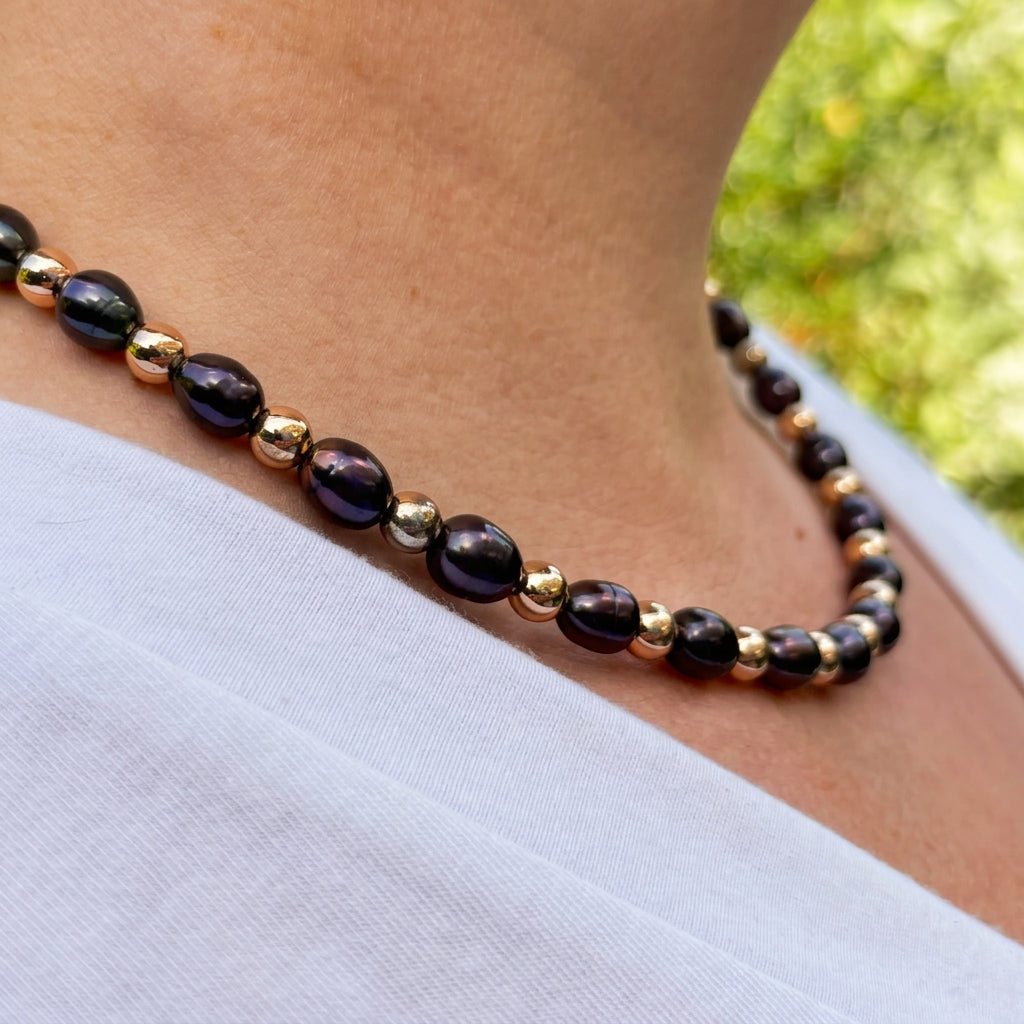 Angled close-up of a deep purple rice pearl necklace worn with a white top, showing the pearls’ irregular oval shapes and lustre between polished gold-coloured hematite beads.⠋
