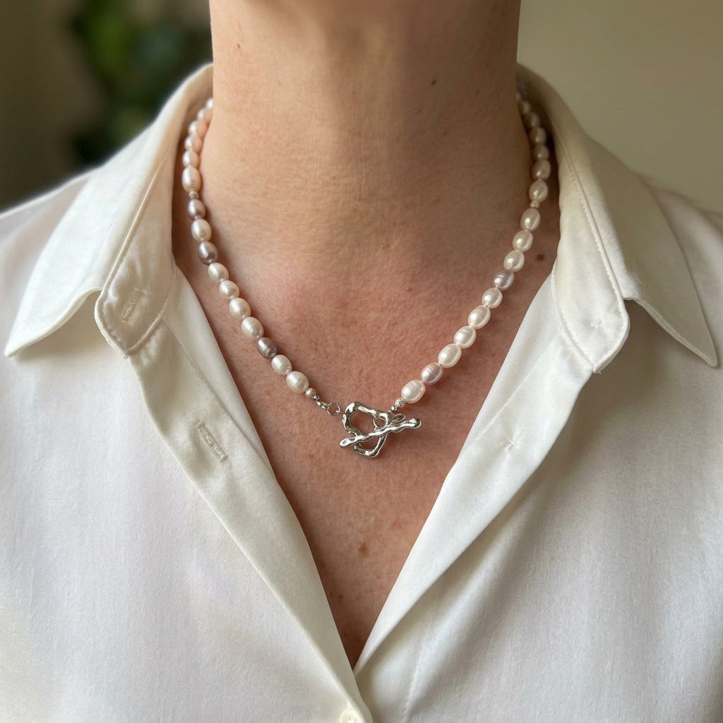 Cream, blush and pale mauve rice pearl necklace with a fastened stainless steel heart-shaped front toggle clasp, worn with an open-neck white shirt.