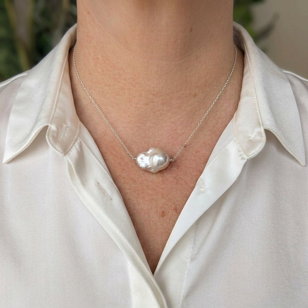 Large blush-pink keshi pearl on a delicate chain, worn with an open-neck white shirt, showing the pearl’s irregular petal-like shape and silvery iridescent lustre.